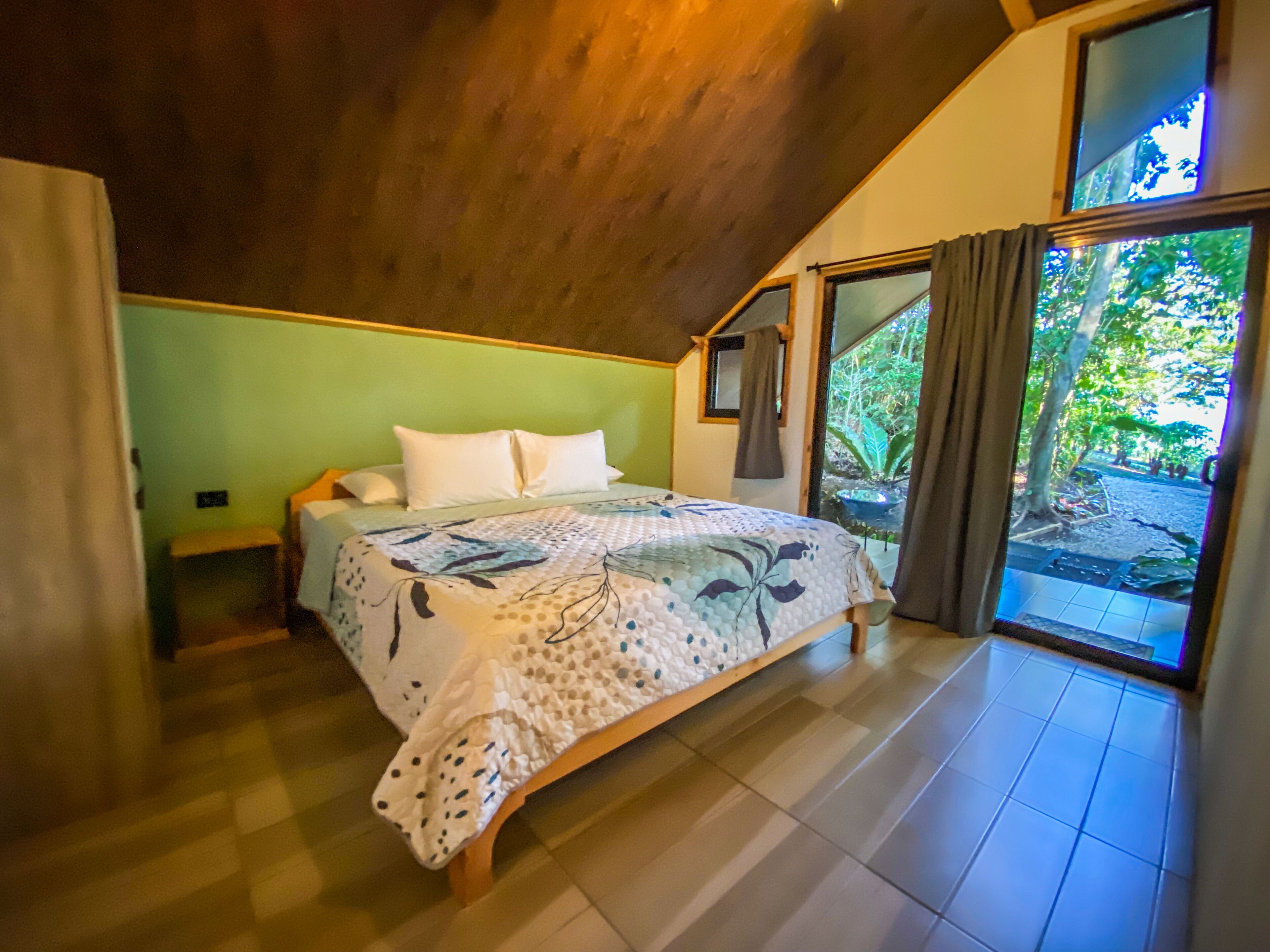Une chambre avec un plafond en bois, un lit recouvert d’une couvre-lit à motifs floraux et une vue sur la verdure, visible à travers une porte vitrée.