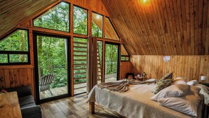 A bedroom with a large bed, wooden walls, and a view of the forest through large windows. - Tityra Lodge (Monteverde)