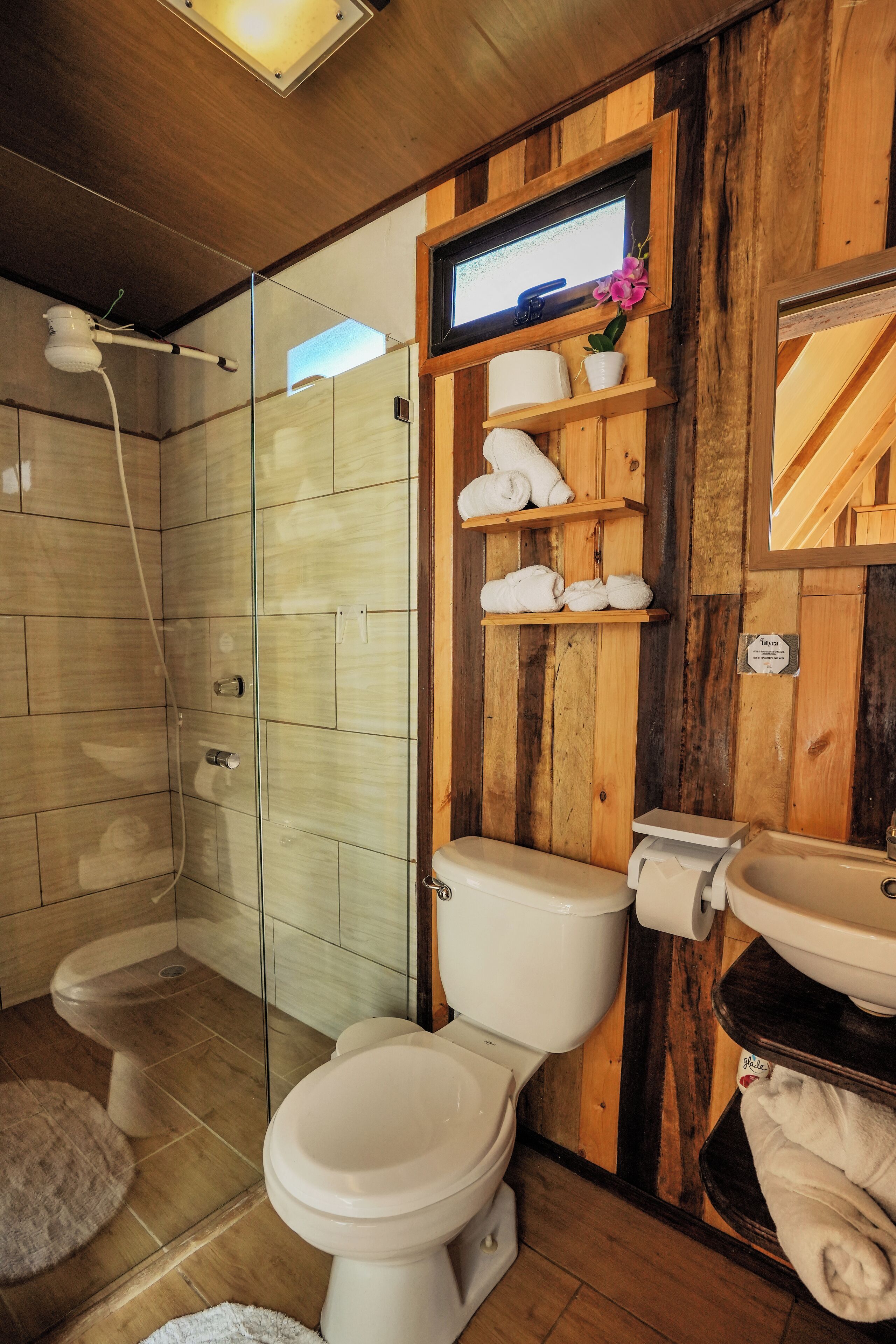 A bathroom with a wooden wall, a glass shower enclosure, a toilet, a sink, and a shelf with towels.