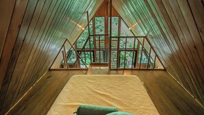 A wooden interior with a bed, pillows, and a view of trees through a window. - Tityra Lodge (Monteverde)