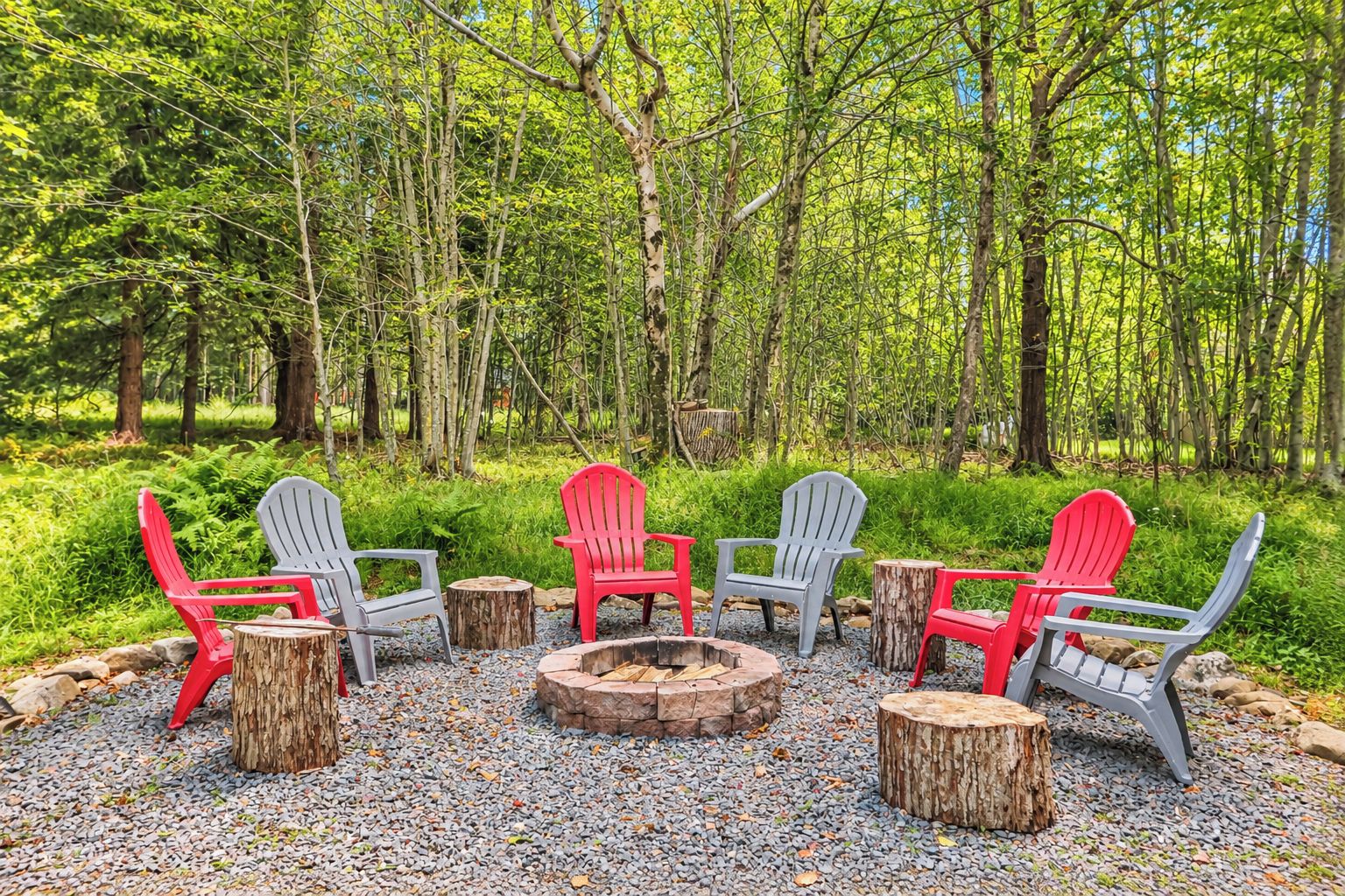 A forest setting with a fire pit and Adirondack chairs.