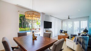 A modern living room with a wooden dining table, comfortable seating, a flat-screen TV, and a large window with a view of greenery. - Peaceful Oasis in Romantic Zone w/ Pool & Parking (Puerto Vallarta)