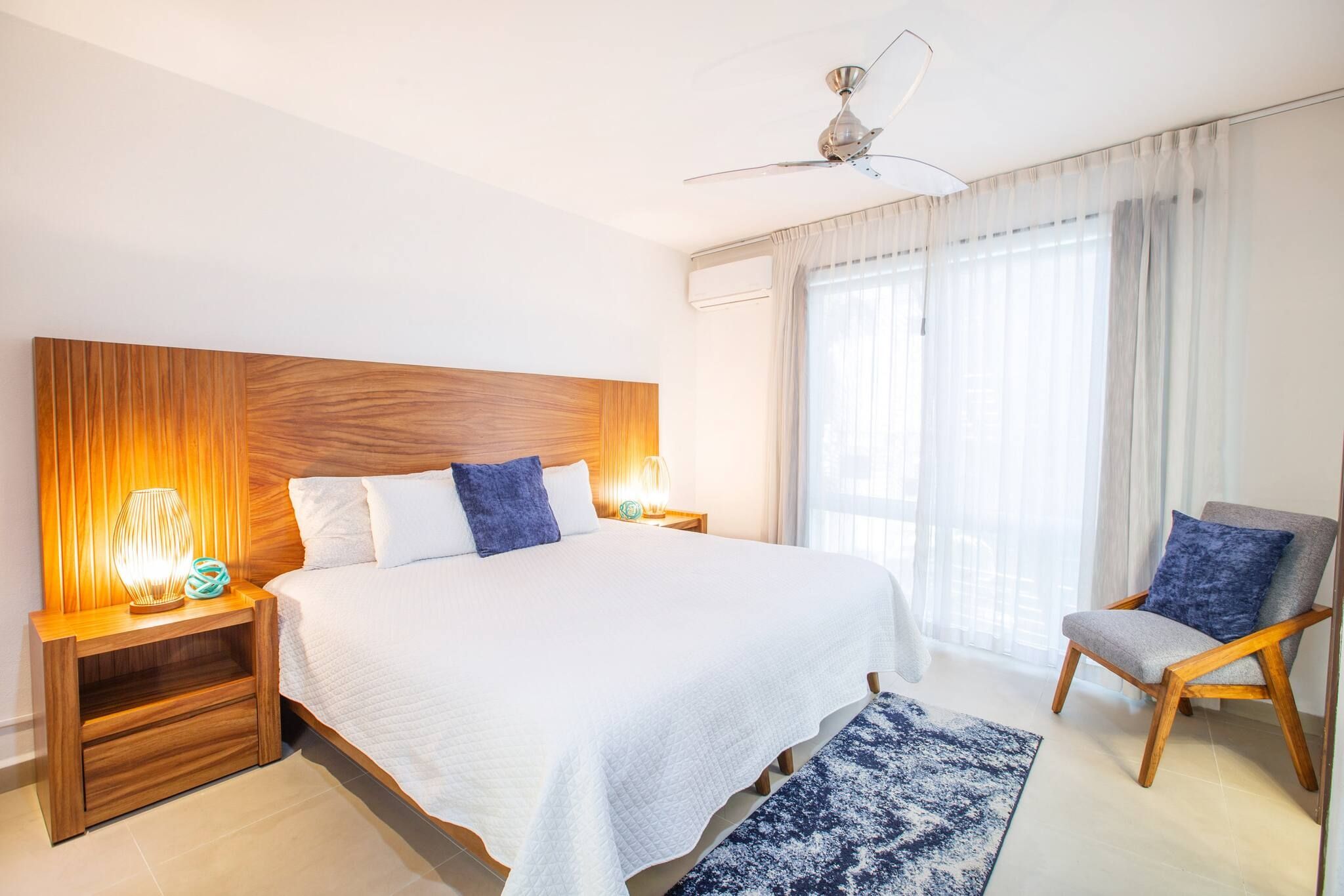 Een hotelkamer met een groot bed, een houten hoofdbord, twee nachtkastjes met lampen, een stoel en een groot raam met gordijnen.