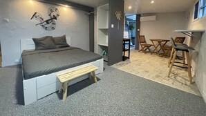 Un dormitorio con cama, mesita de noche y un pequeño banco de madera. En la habitación contigua hay un escritorio con sillas.