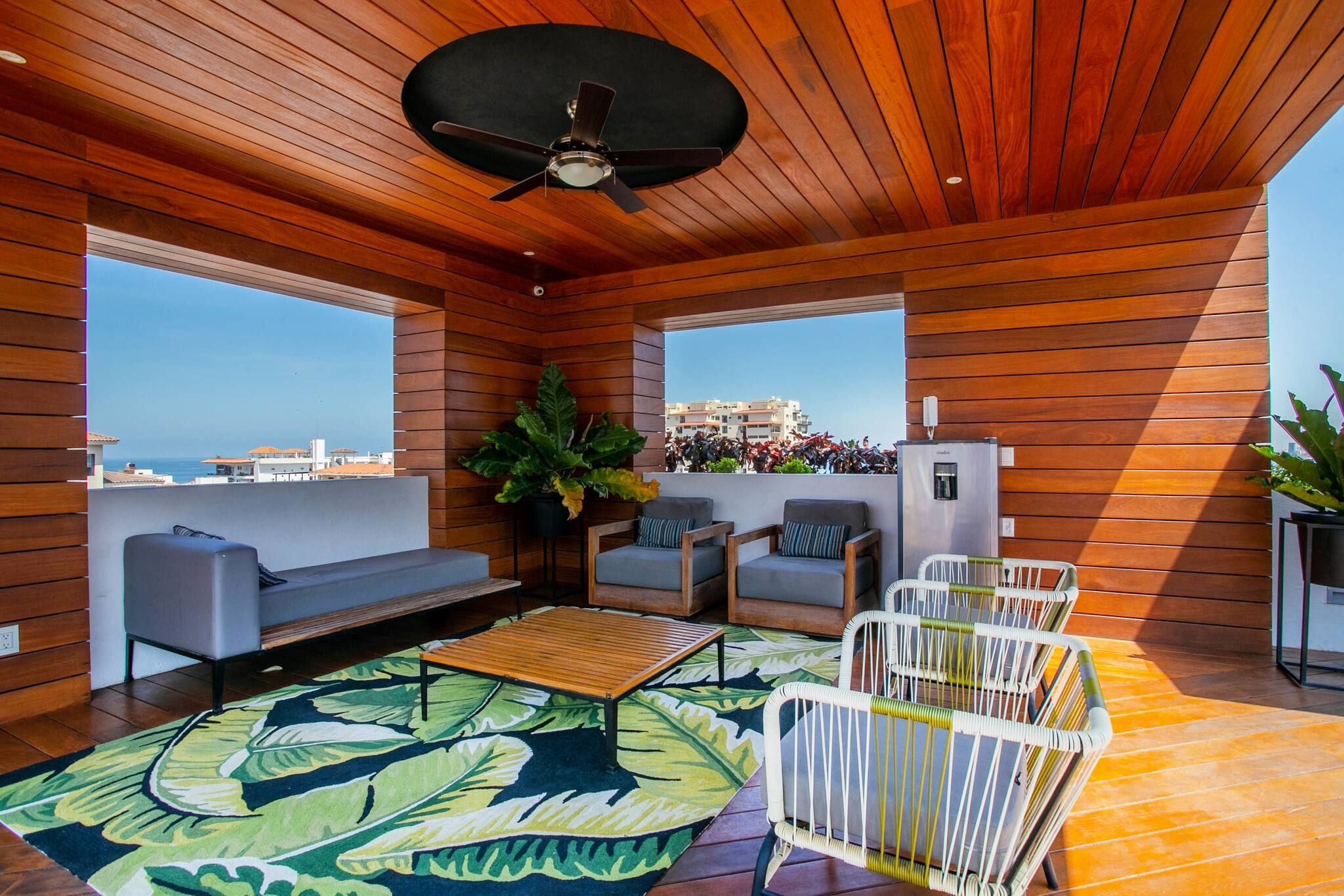 Ein modernes Interieur mit Deckenventilator, Holzwänden, einer Couch, zwei Sesseln, einem Couchtisch und Blick auf die Stadt.