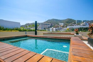 Ein Dachpool mit Holzterrasse, umgeben von Grün und mit Blick auf eine Stadtlandschaft mit Gebäuden und Hügeln.