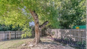 A tree-lined backyard with a wooden fence, a paved walkway, and a green container in the background. - No Cleaning Fee!!~King Bed~Near Dickies,TCU, cultural district~Sleeps 6 (Fort Worth)