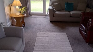 Een woonkamer met een grijze bank, een rode fauteuil, een klein houten tafeltje met een lamp en een groot grijs vloerkleed.
