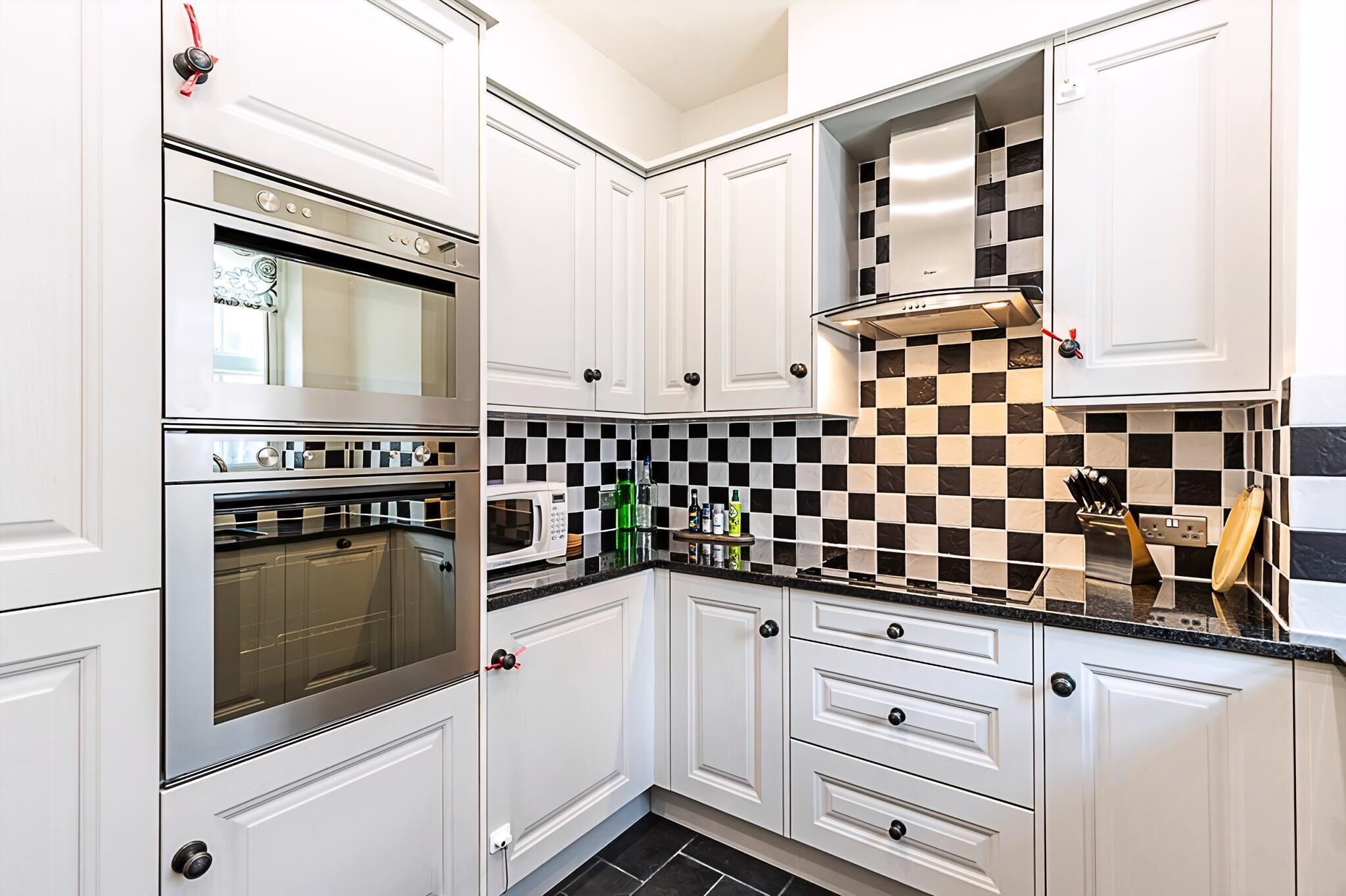 A kitchen with white cabinets, a double oven, a microwave, and a checkered backsplash.
