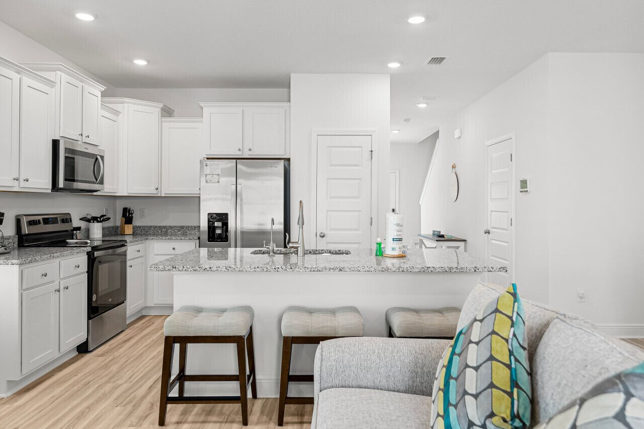 A modern kitchen with white cabinets, a granite countertop, a stainless steel refrigerator, and a built-in microwave. There are three bar stools and a gray sofa with a patterned throw pillow.