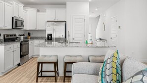 A modern kitchen with white cabinets, a granite countertop, a stainless steel refrigerator, and a built-in microwave. There are three bar stools and a gray sofa with a patterned throw pillow. - NEW! Townhome in Beautiful Santa Rosa Beach! (Santa Rosa Beach)