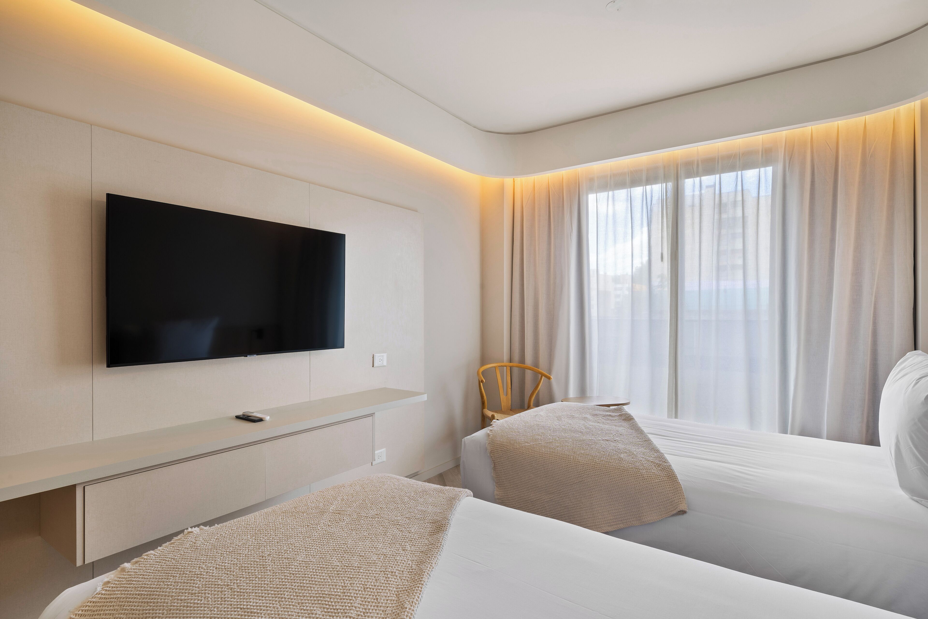 A modern hotel room with a flat-screen TV mounted on the wall, two beds with white linens, a large window with sheer curtains, and a wooden chair.