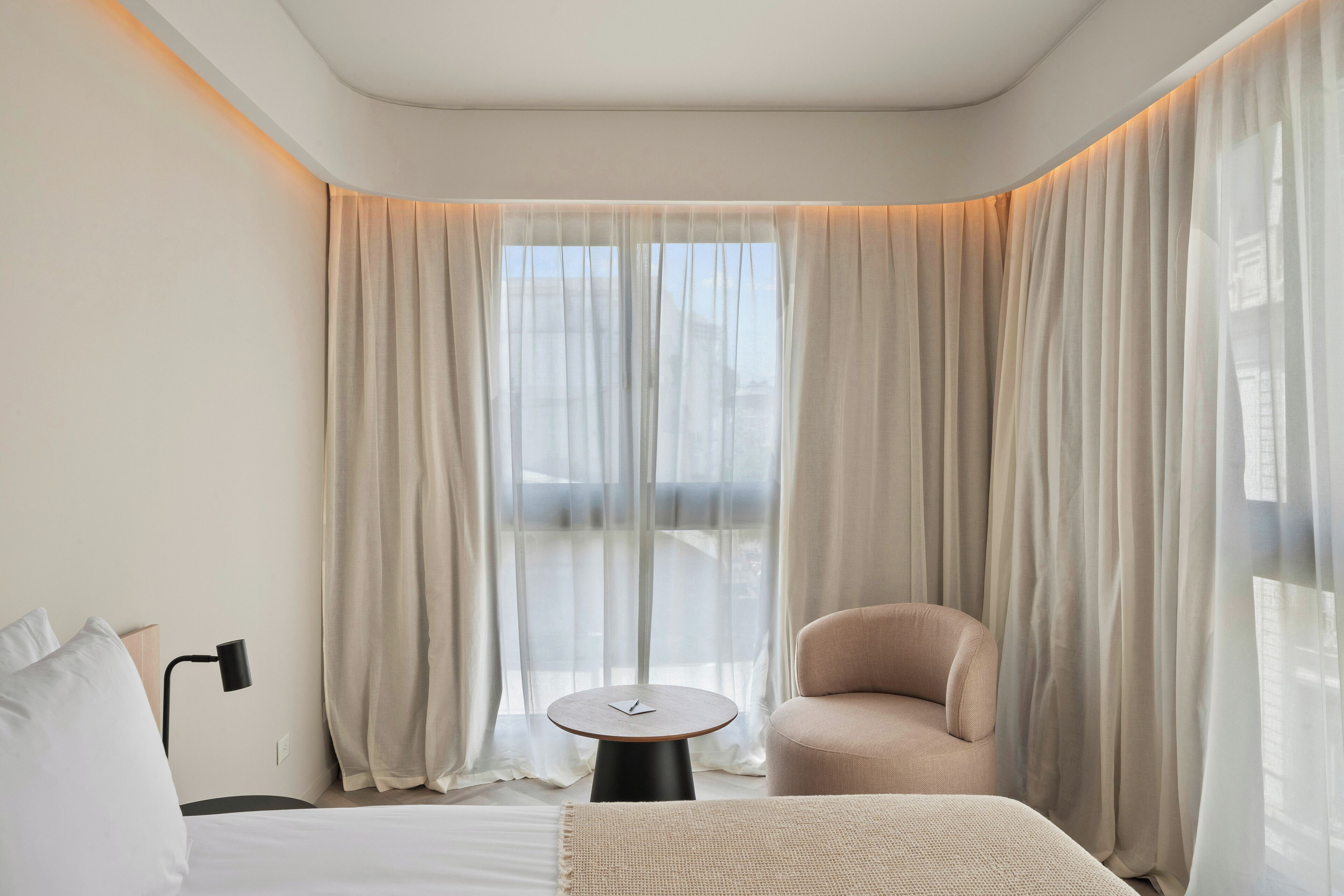 A hotel room with a bed, a small round table, a chair, and sheer curtains.