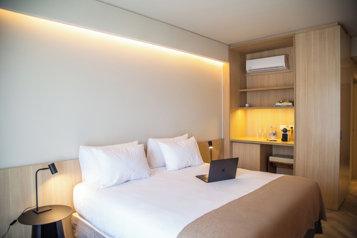 A hotel room with a bed, a laptop, a bedside lamp, a shelf with toiletries, and a wall-mounted air conditioner.
