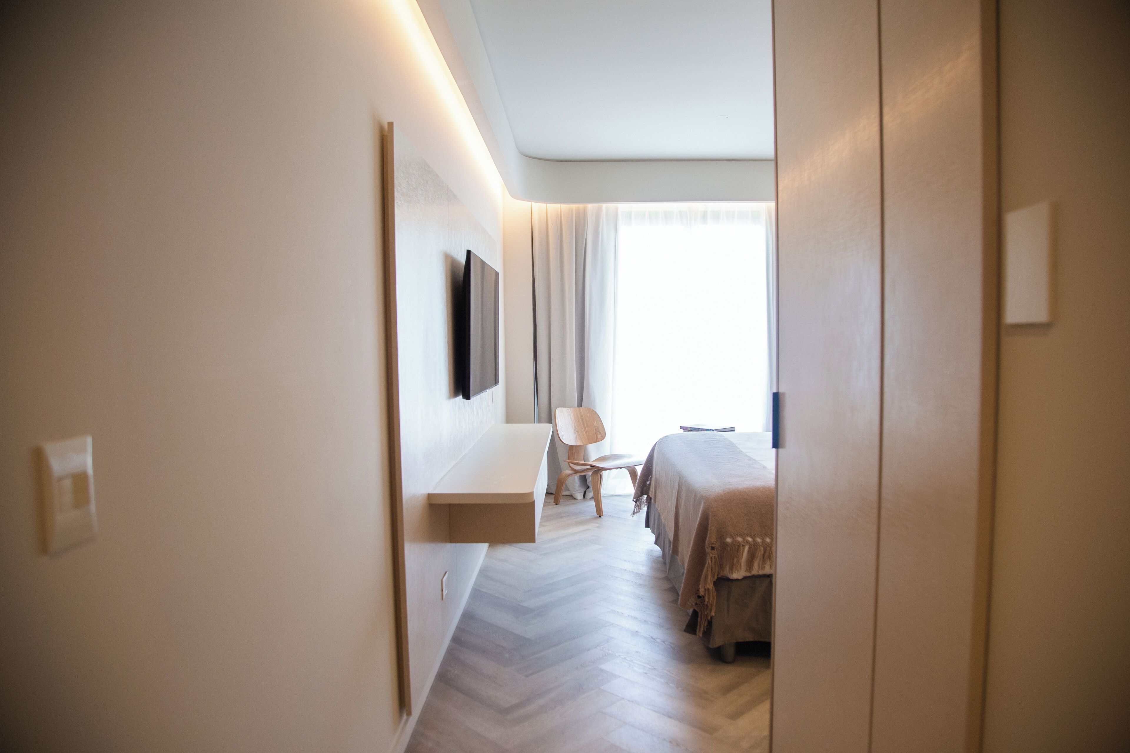 A hotel room with a bed, a chair, a TV mounted on the wall, and a window with curtains.