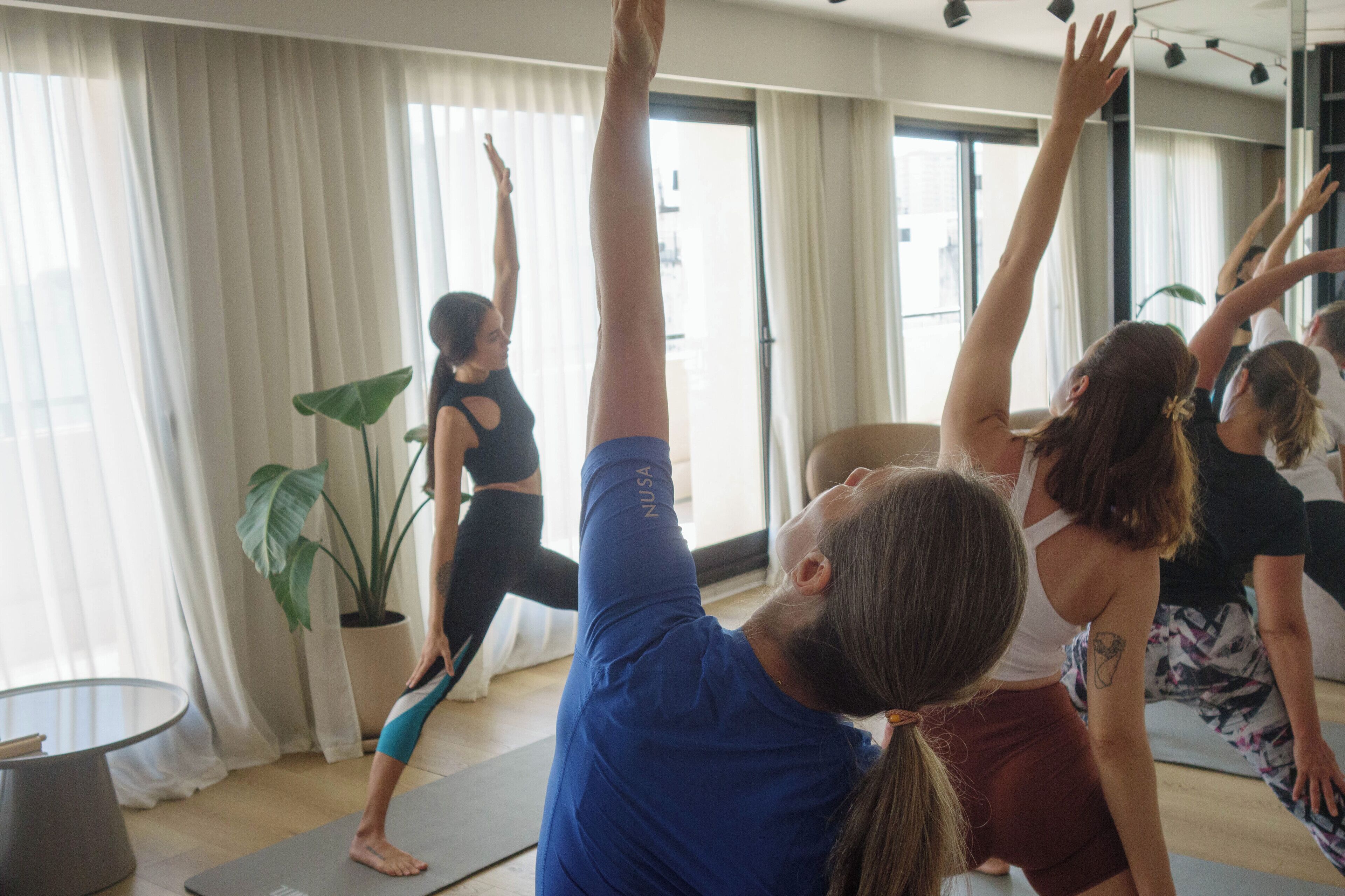 Un cours de yoga dans une pièce dotée de grandes fenêtres, d’une plante en pot et d’une table ronde.