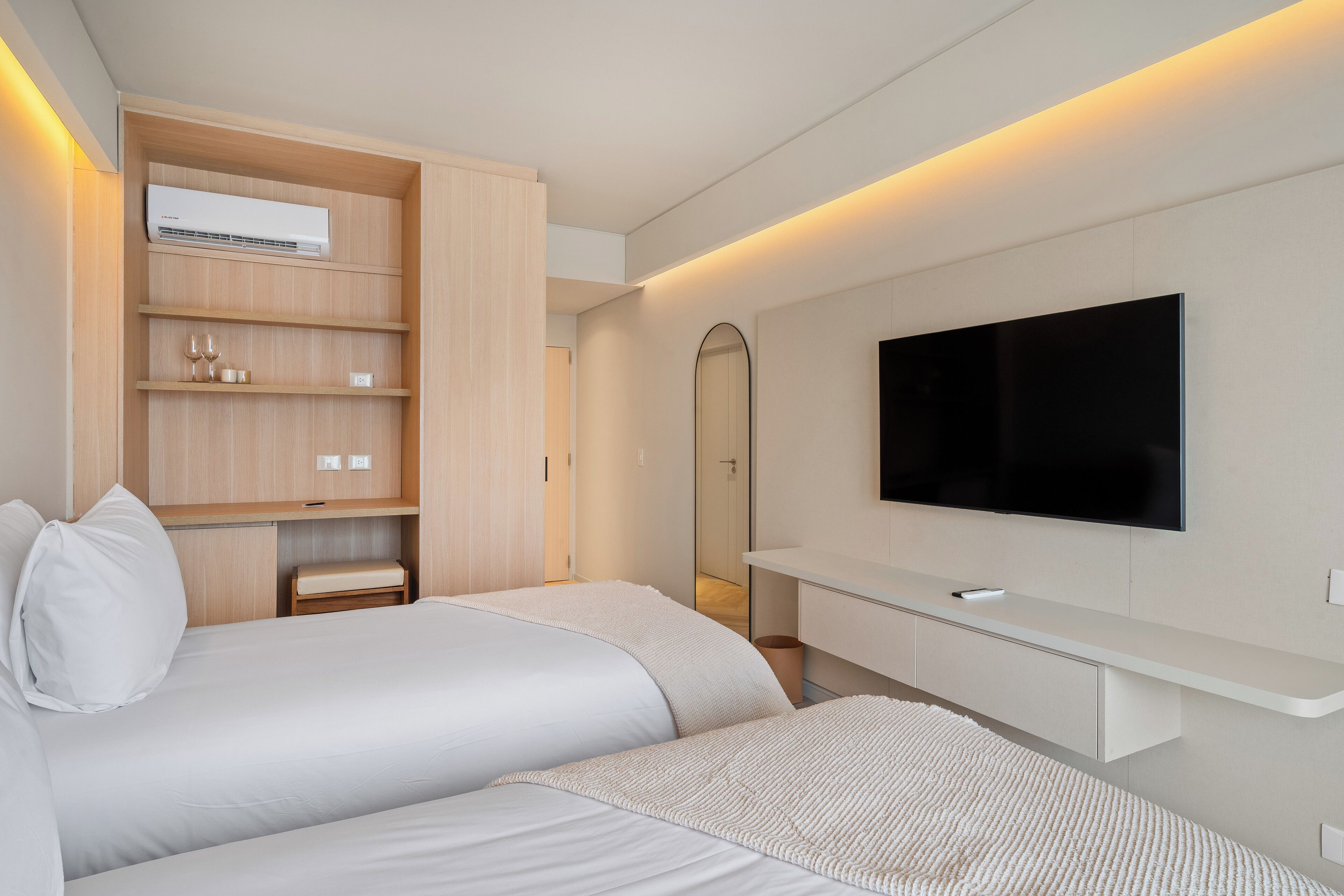 A modern hotel room with two beds, a built-in TV, a mirror, and a wooden shelf unit.