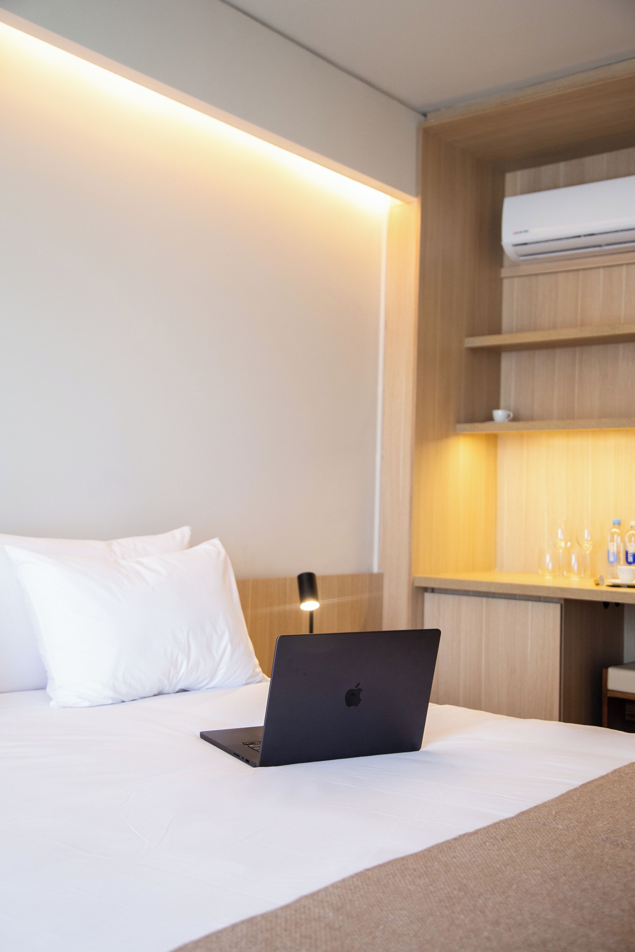 Habitación de hotel con cama, una laptop sobre la cama, una lámpara de noche y un estante de madera con una botella de agua y vasos.