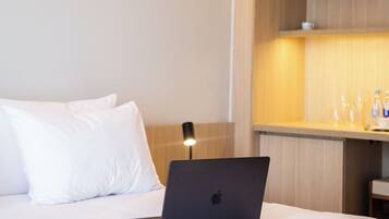 Et hotelværelse med en seng, en bærbar computer på sengen, en sengetavle og en træhylde med en vandflaske og glas.