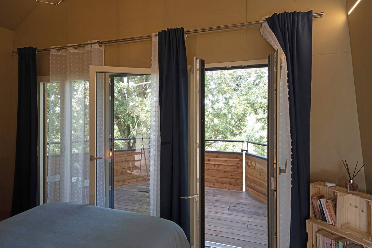 A bedroom with a large bed, a wooden bookshelf, and a view of a wooden deck and trees through open sliding doors.