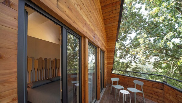 Una terraza de madera con una cama, dos sillas, ventanas amplias y vistas a árboles.