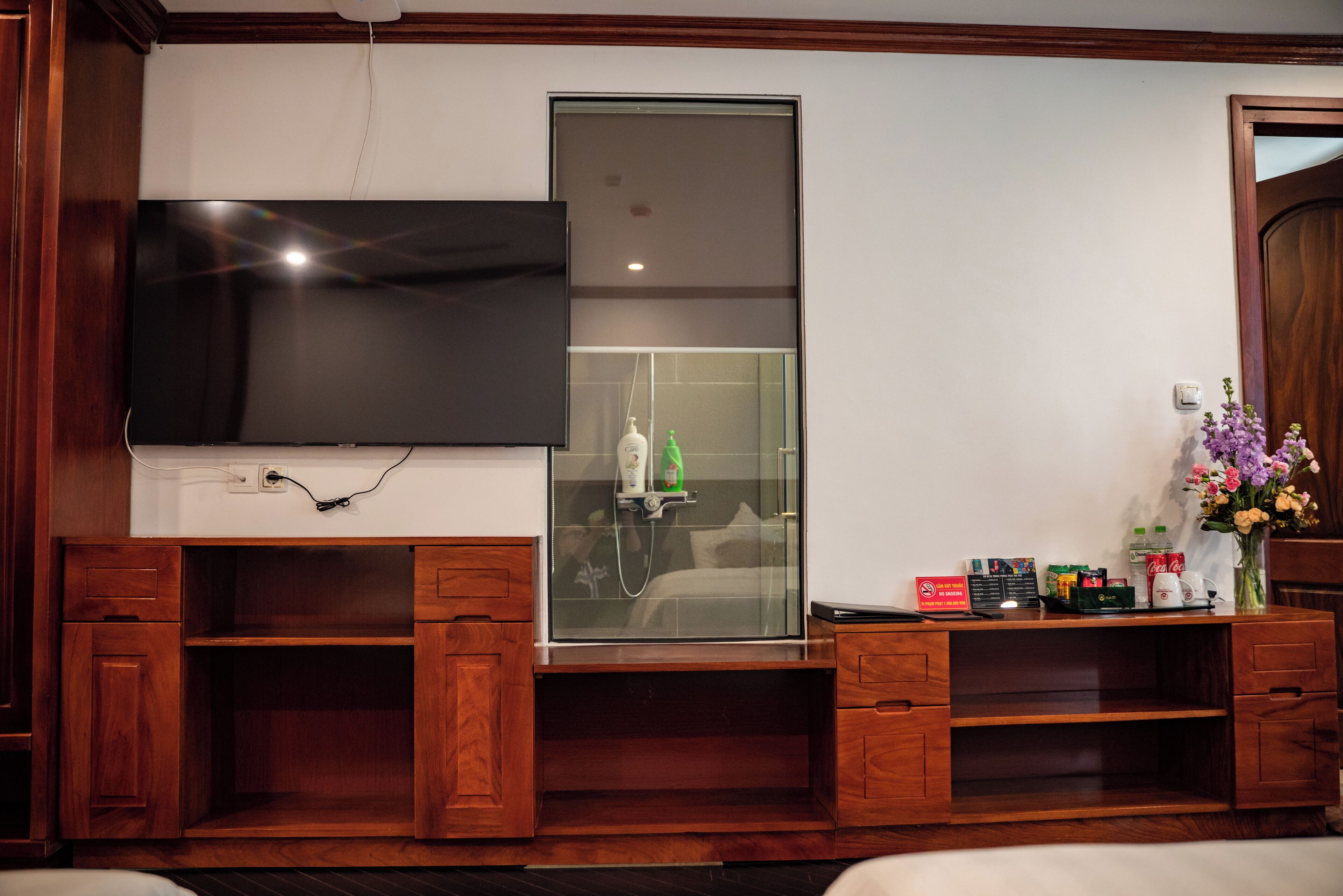 A room with a flat-screen TV mounted on the wall, a glass shower enclosure, wooden cabinets, and a small table with toiletries.