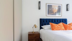 Een slaapkamer met een blauw gestoffeerd hoofdeinde, een houten nachtkastje met een lamp, en een lijst met een afbeelding aan de muur.