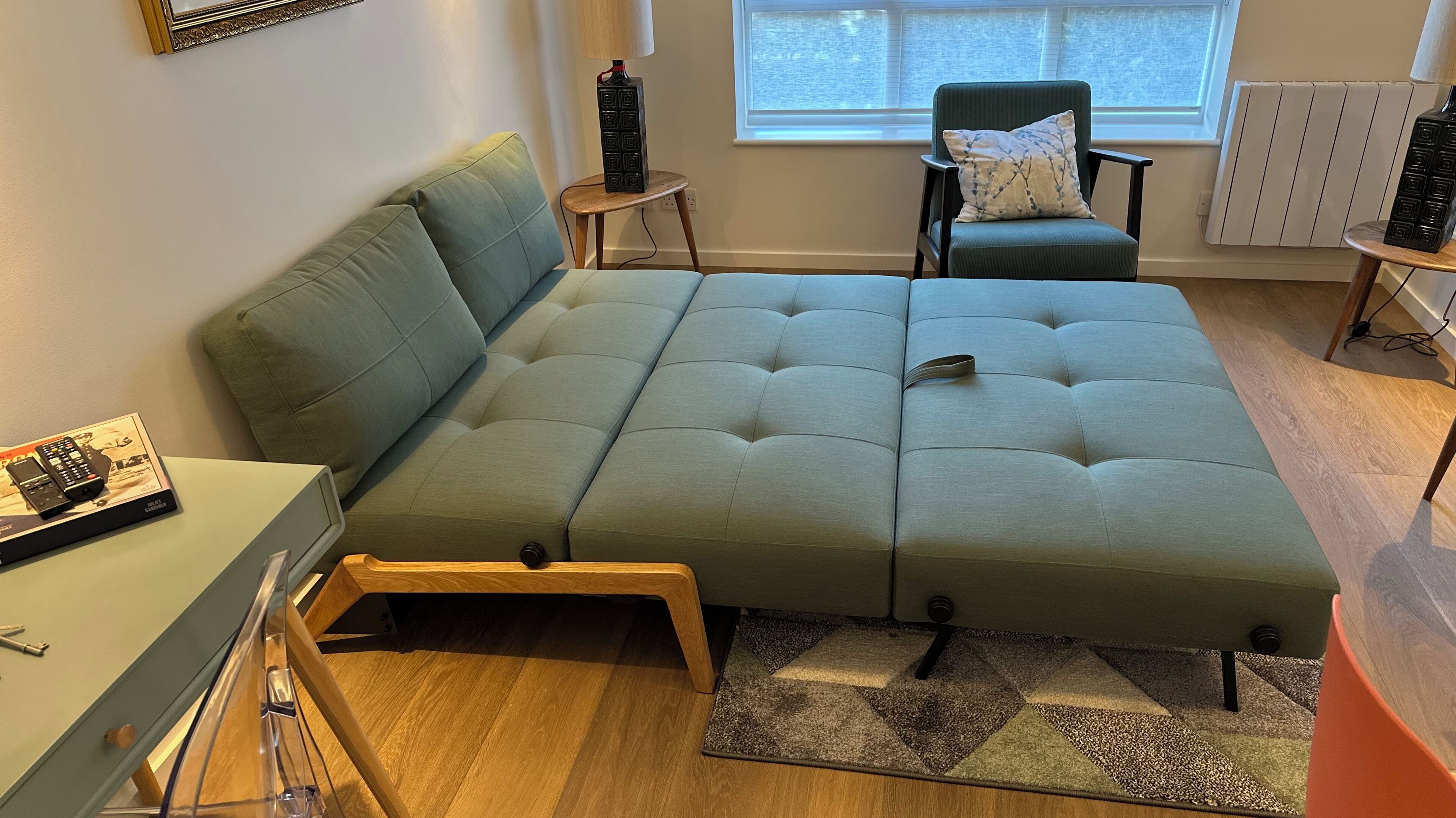 Un espacio habitable moderno y funcional con sofá cama, una mesita auxiliar, una silla y una ventana que proporciona luz natural.