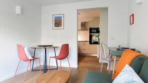 Een moderne woonkamer met een groene bank, twee rode stoelen, een houten salontafel en een eettafel met een lamp. Achtergrond toont een keuken.