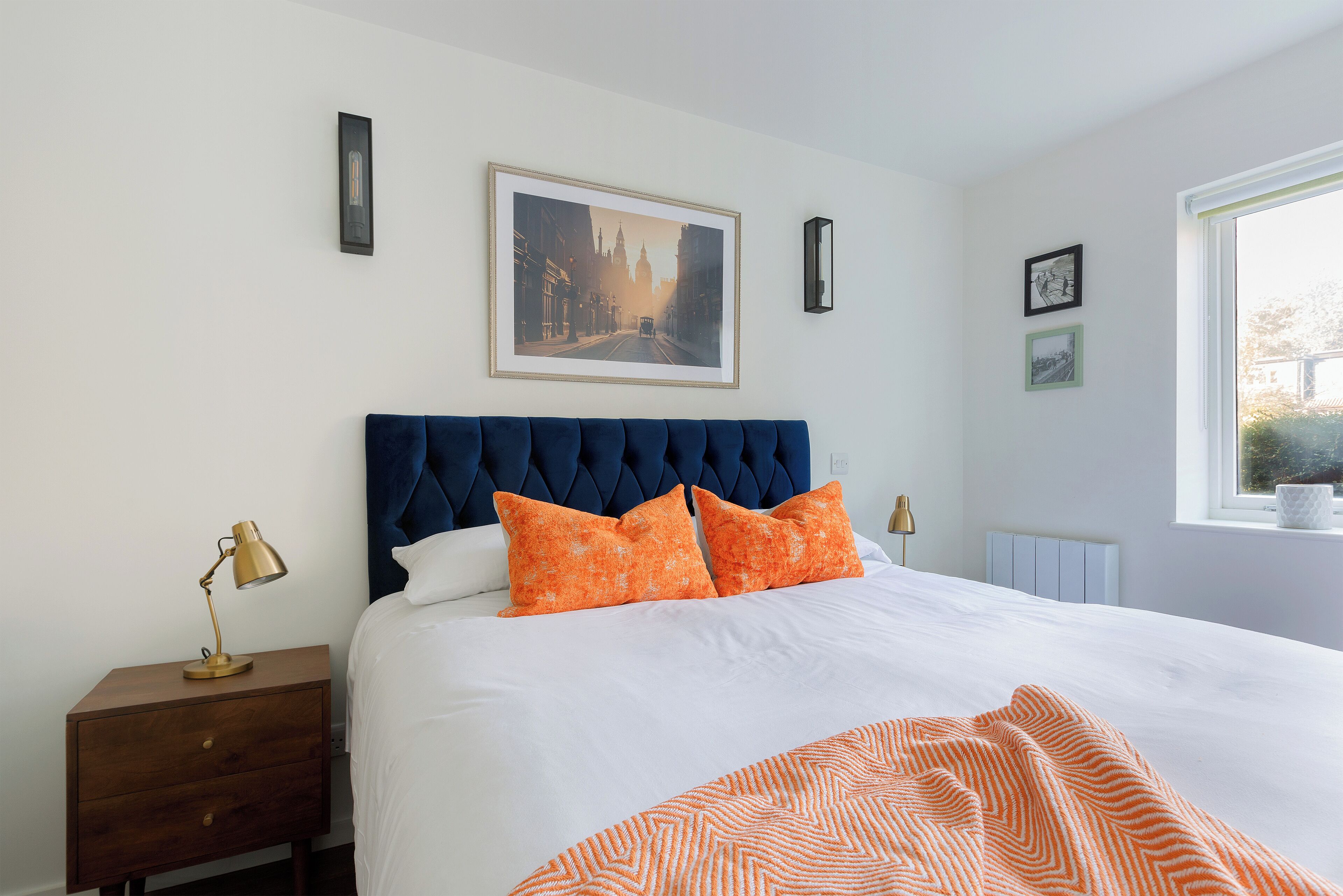 A neatly made bed with a white duvet and orange pillows, a dark blue headboard, a bedside table with a lamp, and a framed picture on the wall.