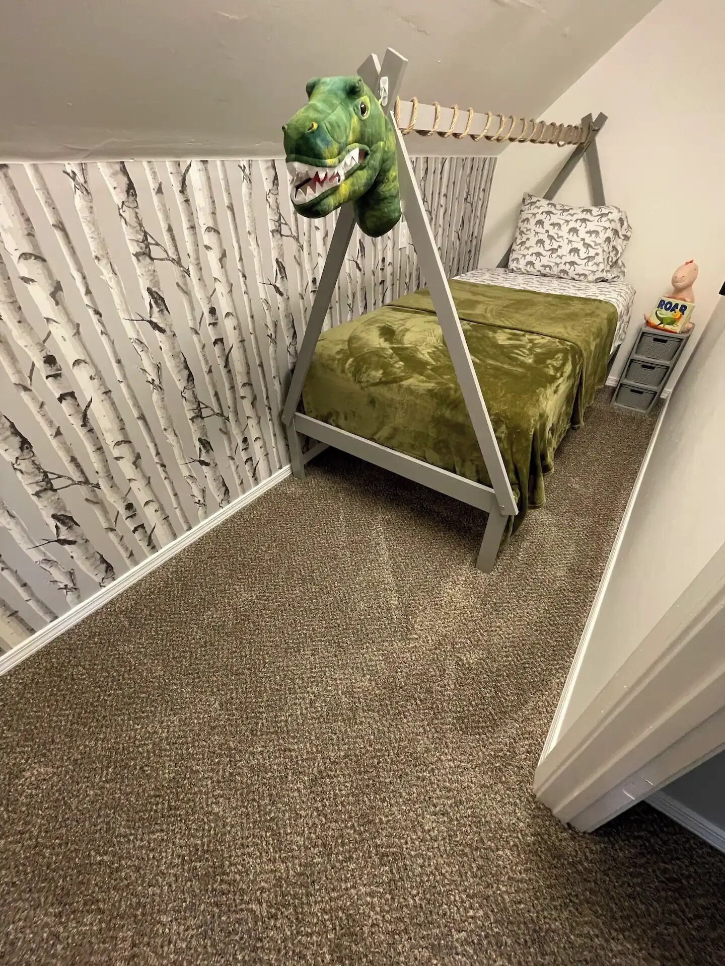 A bedroom with a bed, a green dinosaur head hanging from the ceiling, a patterned wall, and a small nightstand with a toy.