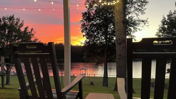 Two wooden Adirondack chairs facing a lake with a sunset in the background.