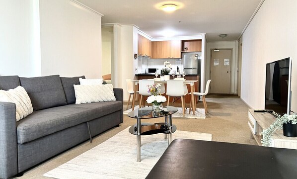 A modern living room with a grey sofa, a coffee table, and a dining area with a white table and chairs. - Spacious City Road Haven (Southbank)