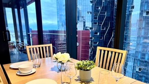 Une table à manger dressée avec des assiettes, des bols, des verres et une plante en pot, dans une pièce moderne dotée de grandes fenêtres et offrant une vue sur la ville.