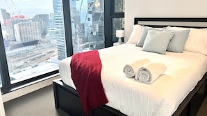 Une chambre d’hôtel moderne avec un grand lit, un plaid rouge et une vue sur le paysage urbain à travers de grandes fenêtres.
