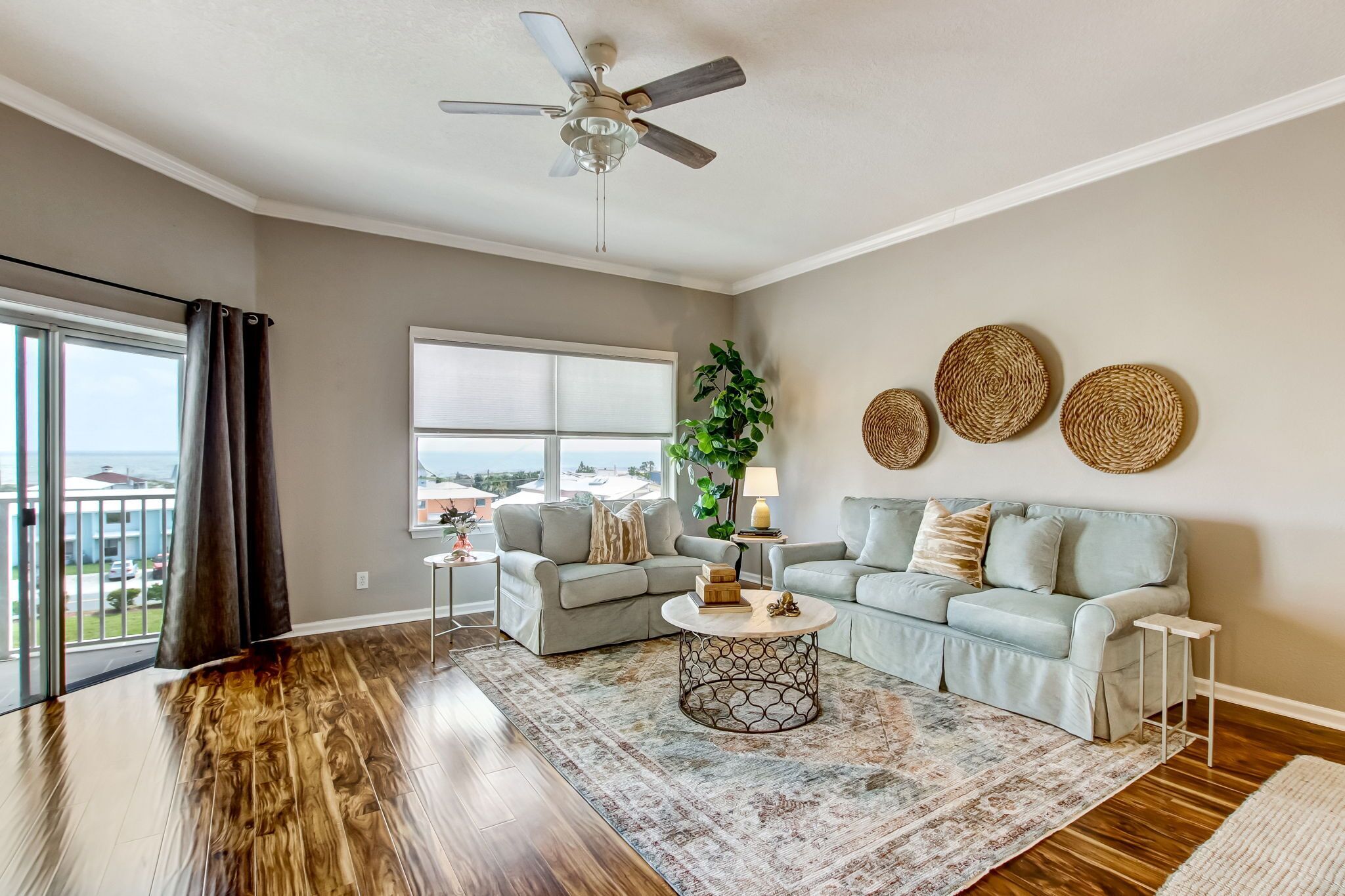 A spacious living room with a large window, a ceiling fan, a grey sofa set, a coffee table, and decorative wall art.