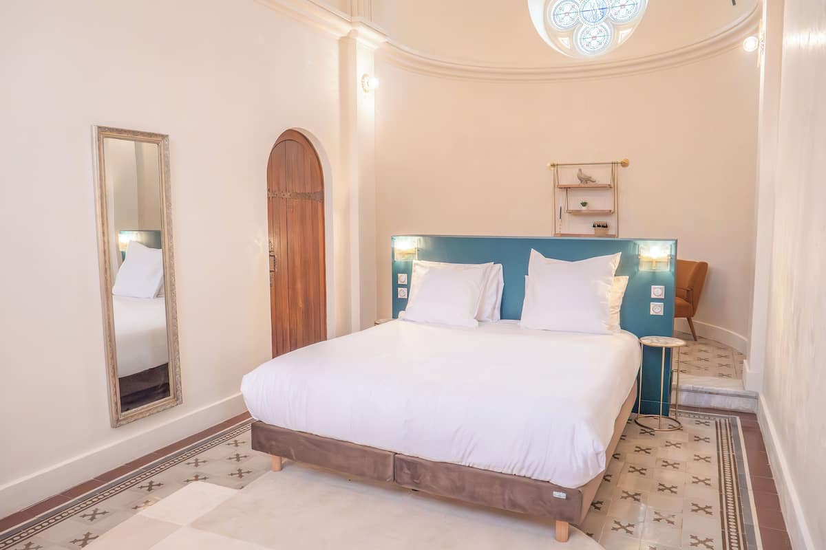 A bedroom with a large bed, a wooden arched door, a mirror, and a small bedside table with a lamp.