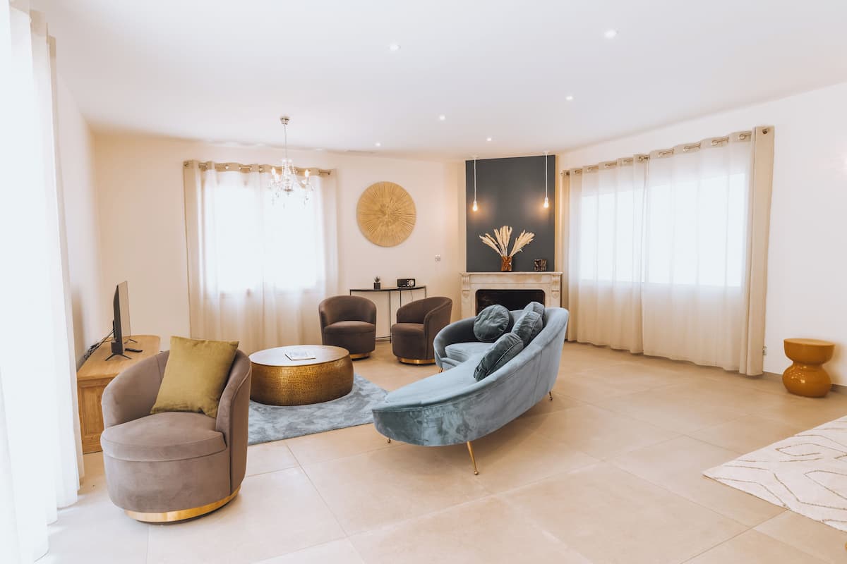 A modern living room with a grey sofa, a round coffee table, and two armchairs. There is a fireplace with a decorative vase and a round mirror above it.