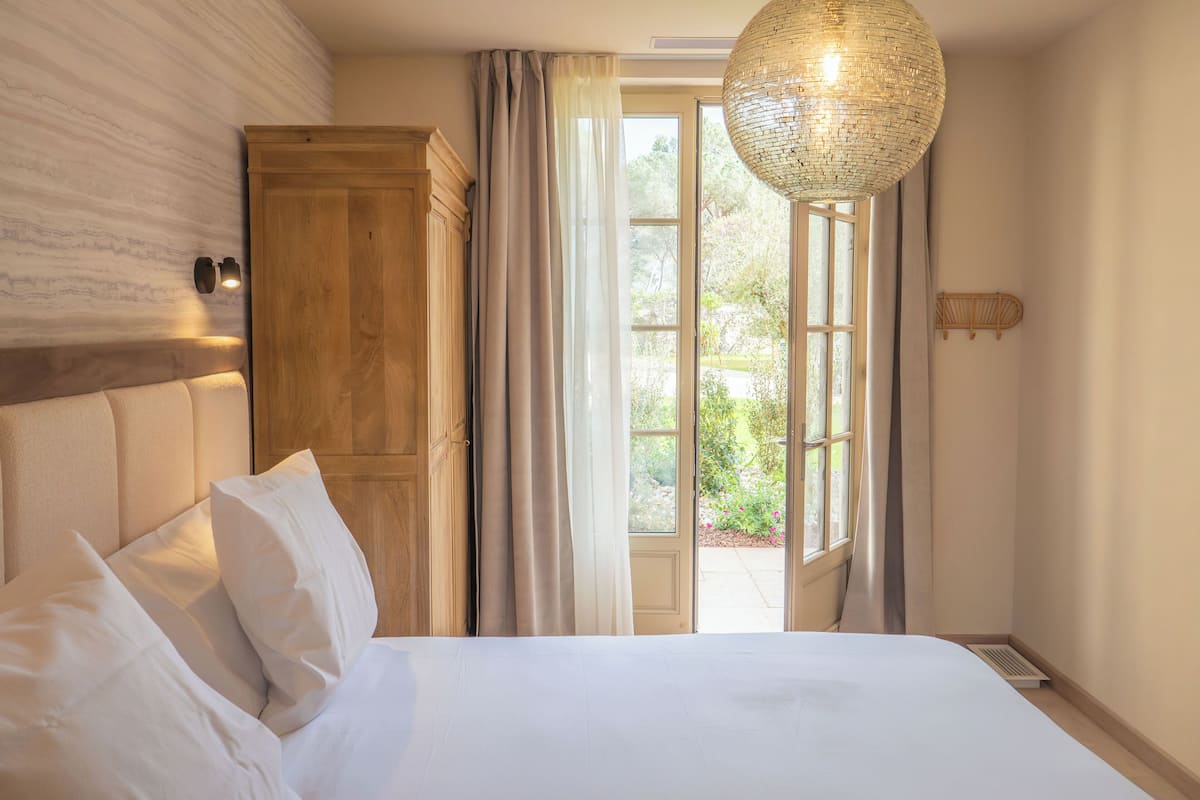 A bedroom with a bed, a wooden wardrobe, a large window with curtains, and a hanging light fixture.