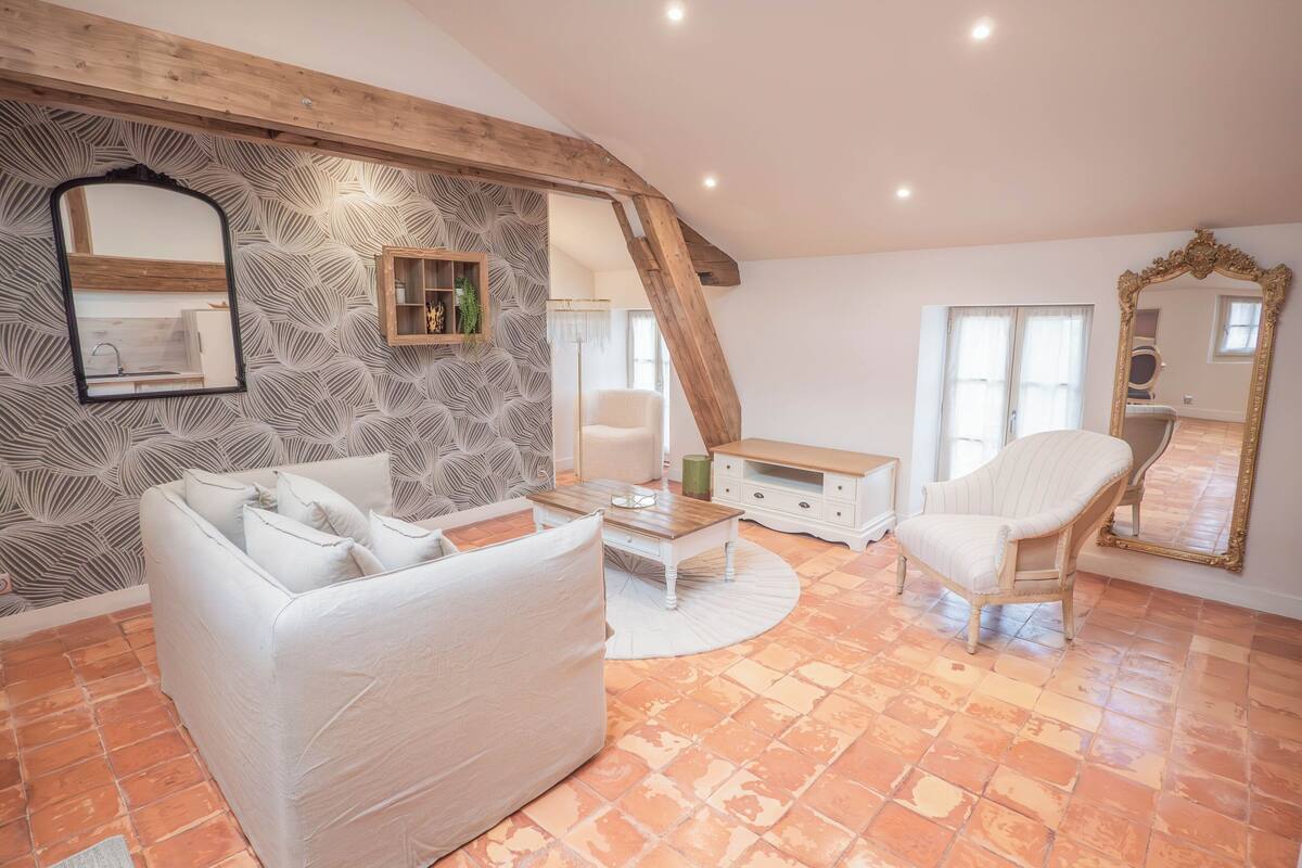 A spacious living room with a white sofa, a coffee table, and a chair. The room features exposed wooden beams, a patterned wallpaper, and a large mirror.