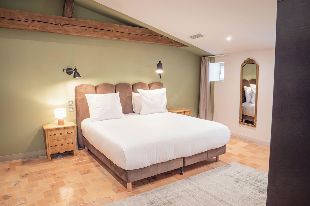 A bedroom with a large bed, wooden headboard, bedside lamps, a mirror, and a tiled floor.