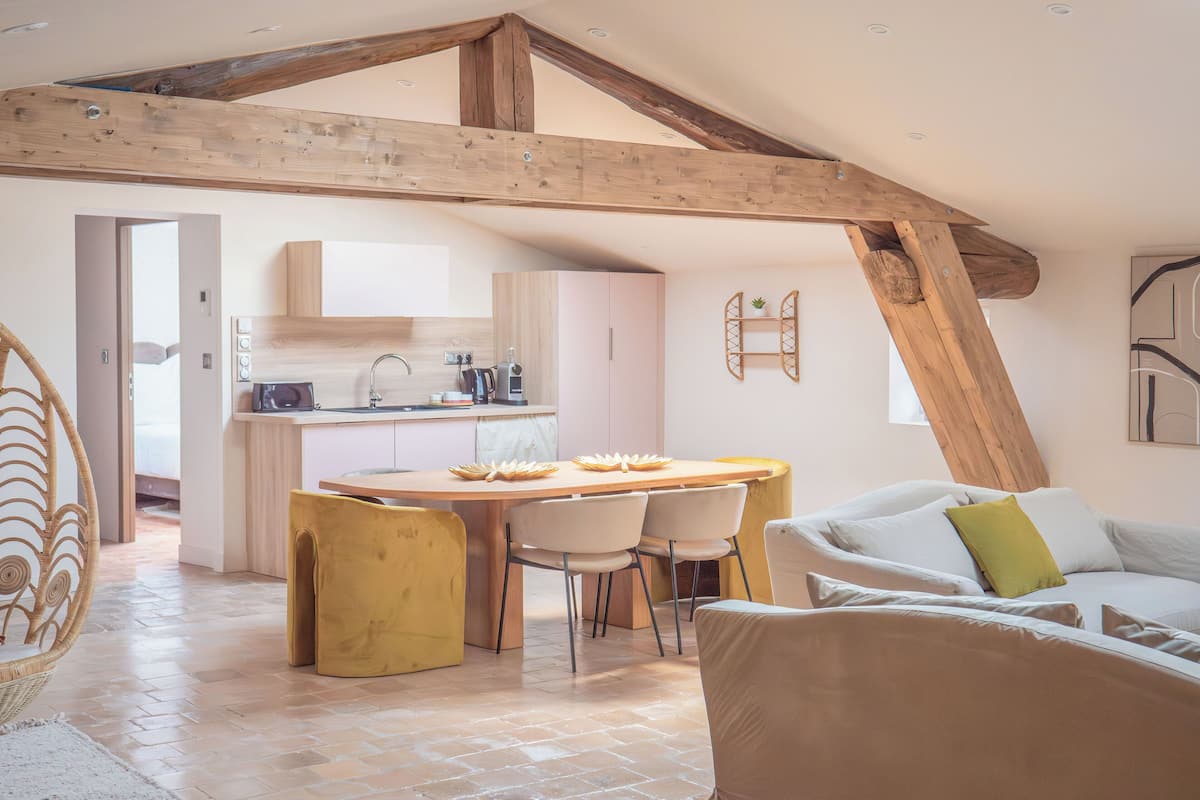 A spacious loft with exposed wooden beams, a kitchen with modern appliances, a dining area with a wooden table and chairs, and a cozy seating area with a sofa and cushions.