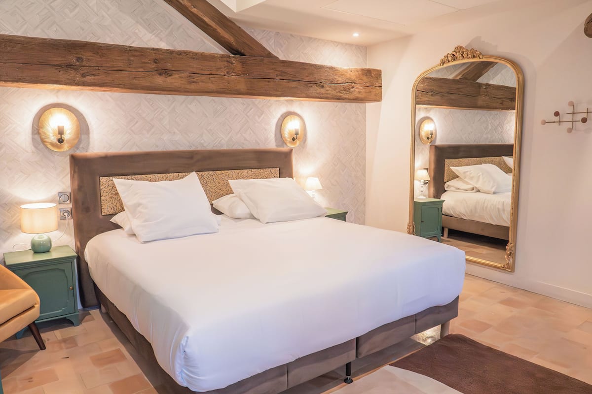 A neatly made bed with white linens and a wooden headboard, flanked by two bedside tables with lamps, in a room with exposed wooden beams and a large mirror on the wall.