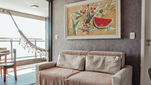 A modern living room with a beige sofa, a hammock, and a large painting of fruits and vegetables on the wall.