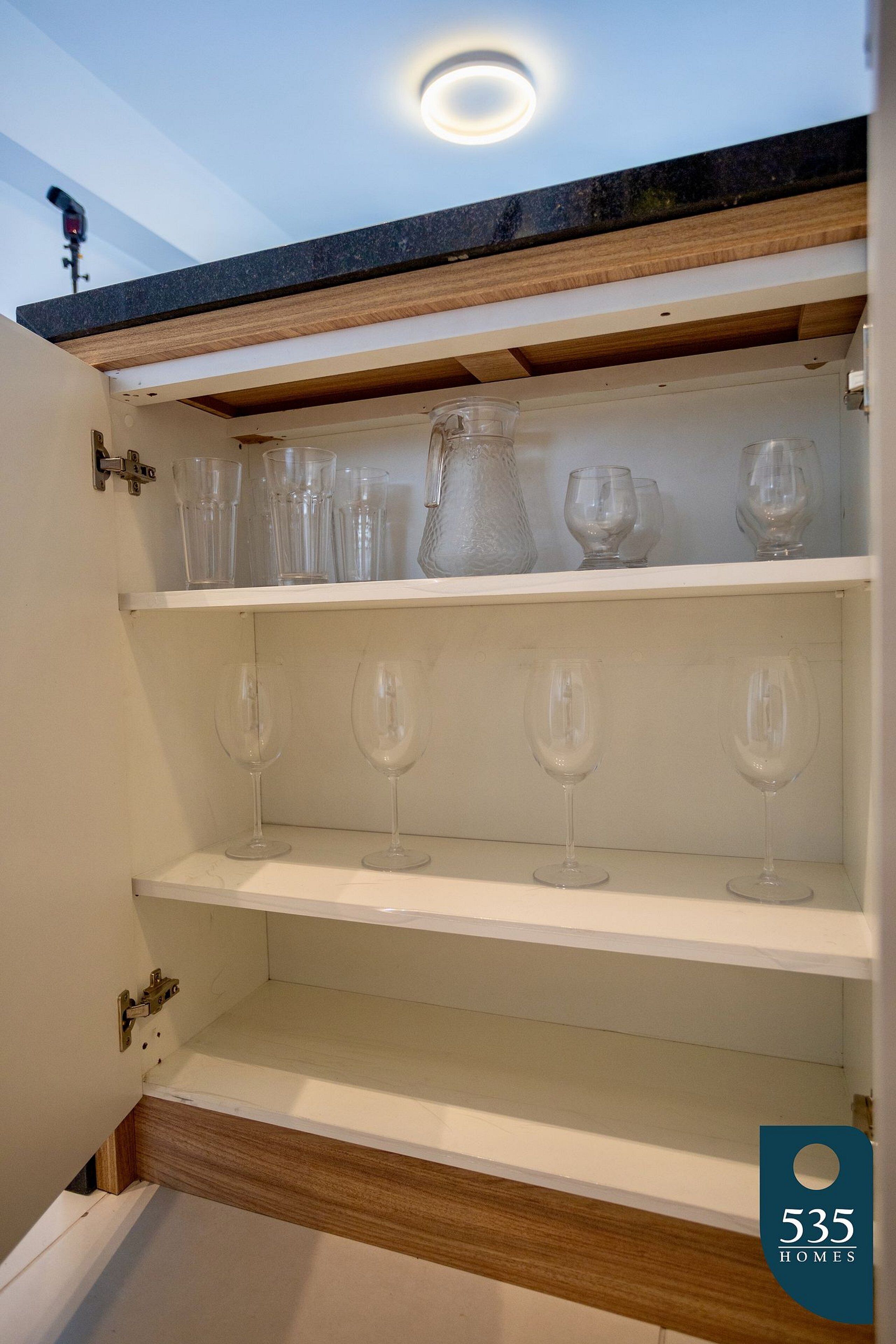 An open kitchen cabinet with shelves holding clear glassware and a pitcher.