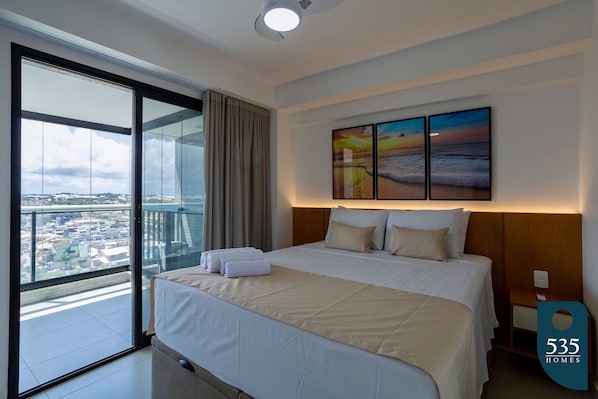 Una habitación de hotel moderna con una cama grande, vistas a la ciudad a través de ventanales del suelo al techo y un cuadro de atardecer en la pared.