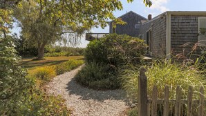 Een grindpad leidt naar een houten chalet met terras, omgeven door weelderige begroeiing en een grote boom.
