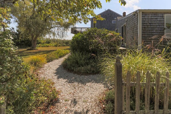 A gravel path leading to a wooden cabin with a deck, surrounded by lush greenery and a large tree. - 8 Sandy Neck- Family Getaway Near Sandy Neck Beach, pet-friendly! (Sandwich)