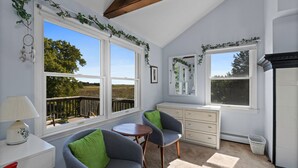 Een gezellige kamer met twee stoelen, een klein tafeltje en een kaptafel. Grote ramen bieden uitzicht op groen en een houten terras daarbuiten.
