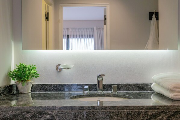Un baño con encimera de granito, lavamanos, espejo, planta en maceta y perchero.